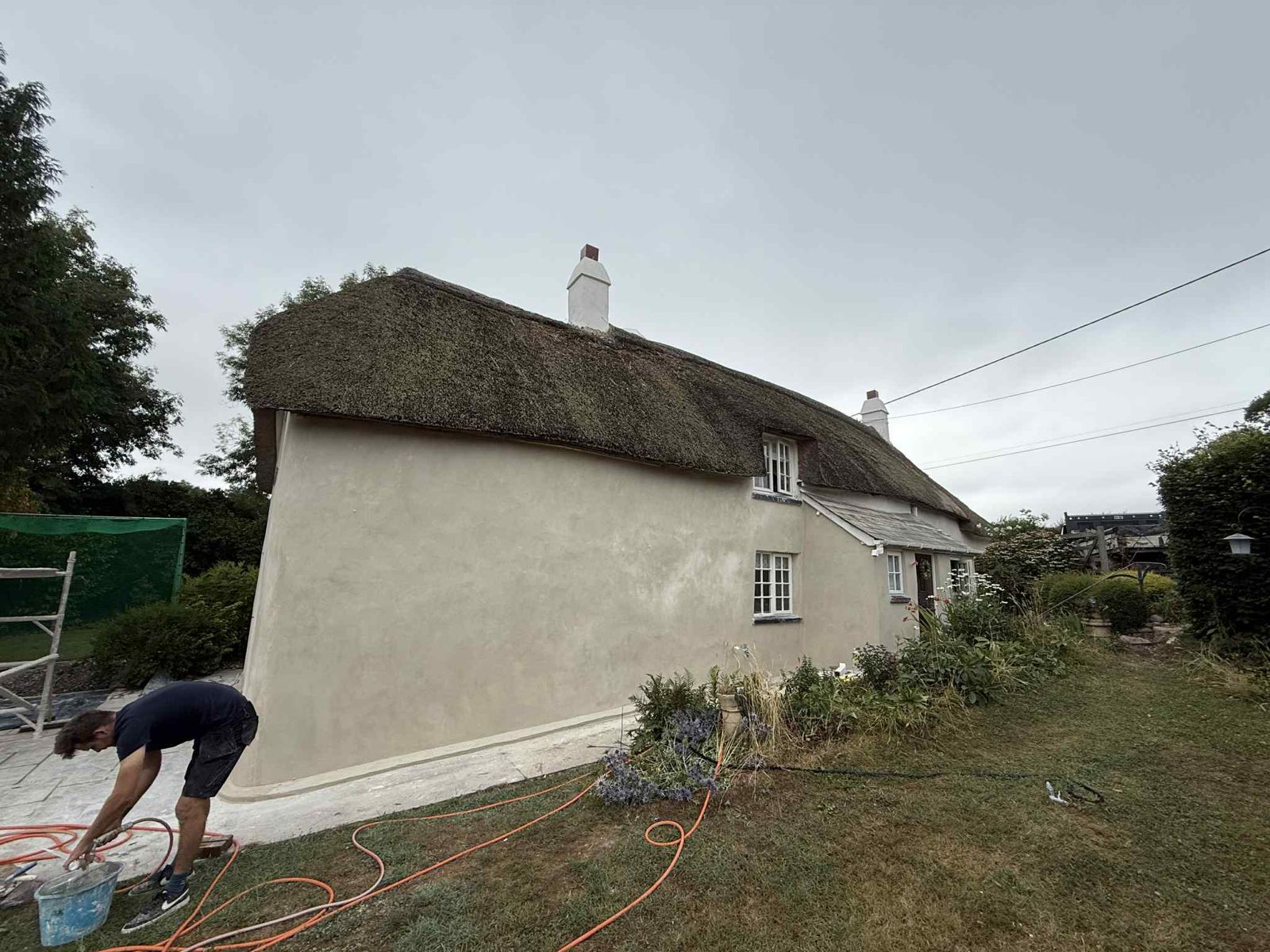 A thatched cottage being rendered, with a plasterer mixing in the garden